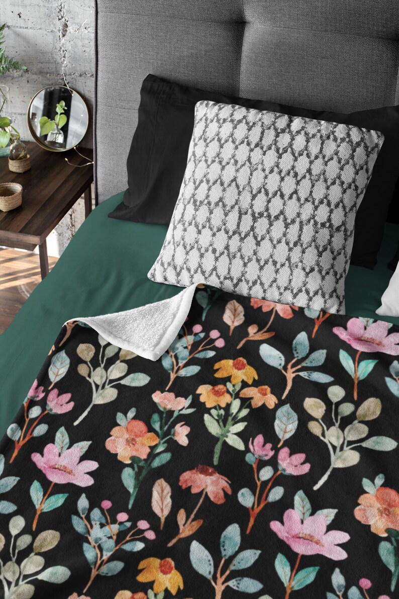 May include: A black blanket with a floral pattern featuring pink, orange, and yellow flowers, along with green and tan leaves. The blanket is on a bed with a white patterned pillow and black pillowcases. The blanket's underside is white.