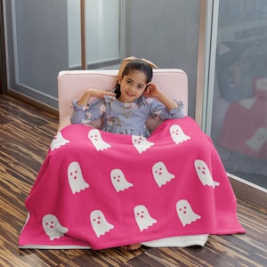 Puede incluir: Una manta de forro polar rosa con un patr&oacute;n de fantasmas blancos. Una ni&ntilde;a est&aacute; sentada en una silla rosa con la manta envuelta alrededor de ella.