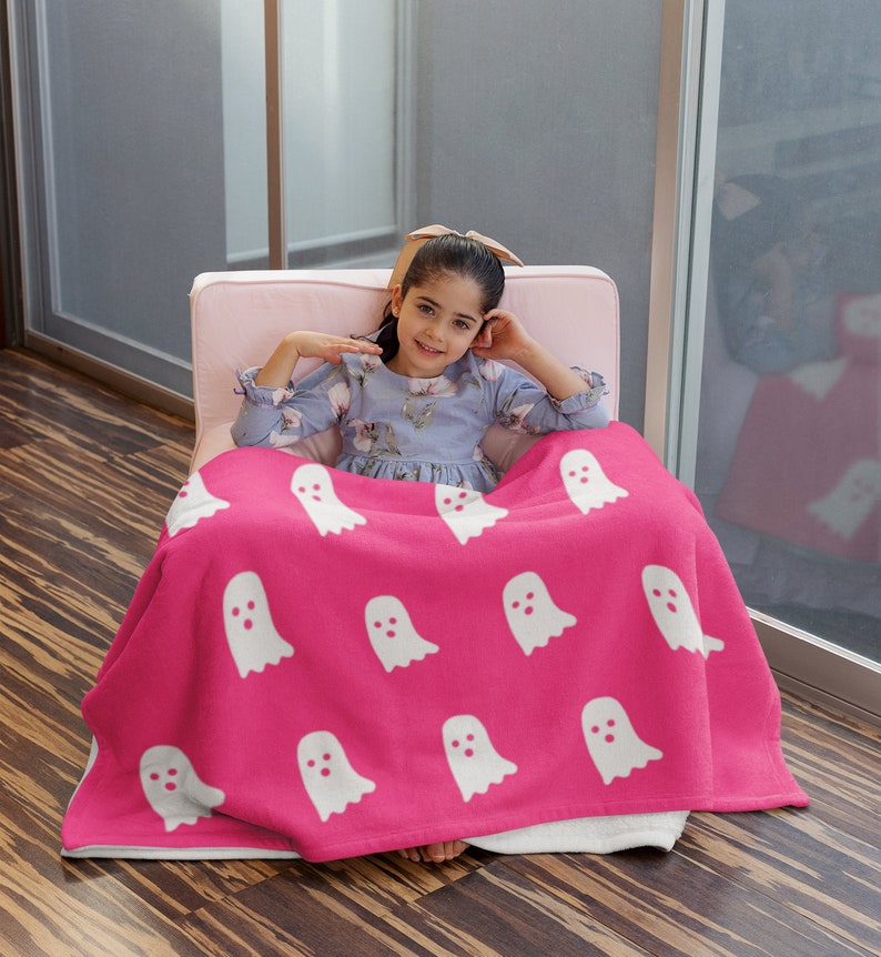 Puede incluir: Una manta de forro polar rosa con un patr&oacute;n de fantasmas blancos. Una ni&ntilde;a est&aacute; sentada en una silla rosa con la manta envuelta alrededor de ella.