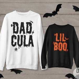 May include: Two Halloween themed sweatshirts. One white sweatshirt with the text "DAD CULA" in black dripping letters. The other black sweatshirt with the text "LIL BOO" in orange dripping letters.