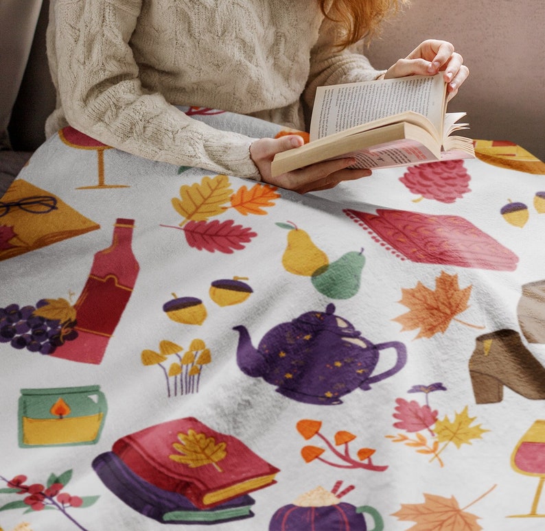Puede incluir: Una manta blanca con un patr&oacute;n de oto&ntilde;o que presenta ilustraciones de libros, copas de vino, bellotas, peras, teteras y hojas.