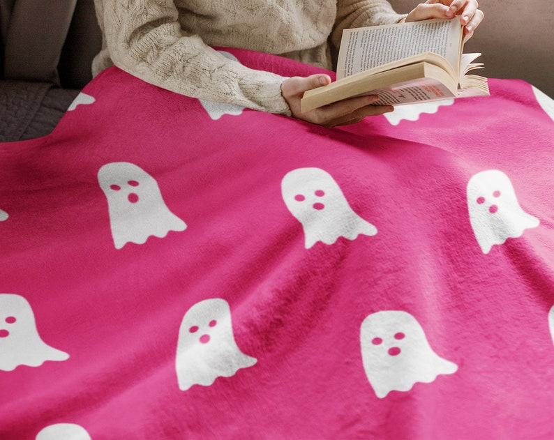 Puede incluir: Una manta de forro polar rosa con un patr&oacute;n de fantasmas blancos. La manta est&aacute; doblada y colocada sobre un sof&aacute;.