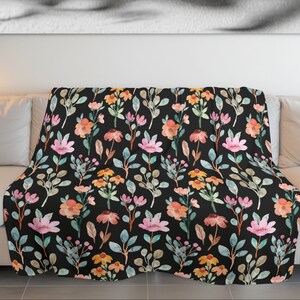 May include: A floral throw blanket with a black background. The blanket features a watercolor pattern of pink, orange, and yellow flowers with green and gray leaves. The blanket is draped over a white couch.