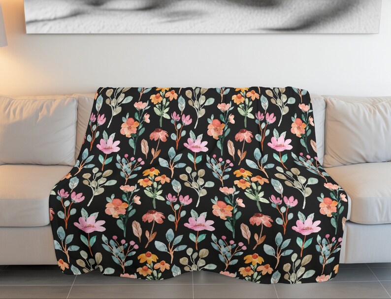 May include: A floral throw blanket with a black background. The blanket features a watercolor pattern of pink, orange, and yellow flowers with green and gray leaves. The blanket is draped over a white couch.