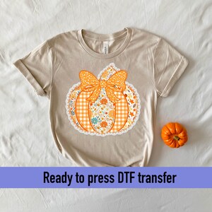 May include: Beige t-shirt with a pumpkin graphic. The pumpkin design features an orange bow, a gingham pattern, and floral details. A small orange pumpkin sits to the right. The text "Ready to press DTF transfer" is at the bottom.