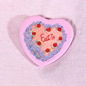 May include: A heart-shaped pink compact mirror featuring a graphic of a cake with light blue frosting, a yellow grid pattern, and eight red cherries. The text "Eat it" is written in red script in the center, with "@shopsoftcore" below.