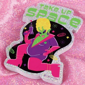 May include: A stack of stickers featuring a green alien character in a purple outfit, holding a ray gun. The stickers have a silver glitter border and the text "TAKE UP SPACE" in green. The background is a pink, glittery fabric.