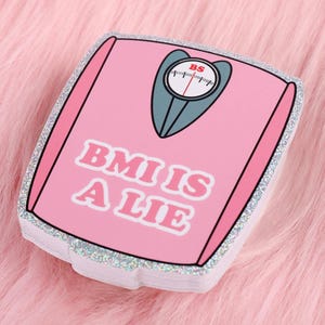 May include: A stack of pink stickers with a scale design and the text "BMI IS A LIE" in white. The scale has a gray face and a red needle. The stickers have a glittery silver border.