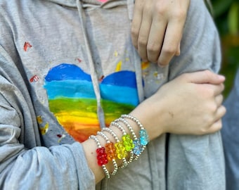 Könnte beinhalten: Ein Set bunter Gummibärchen-Charm-Armbänder. Die Armbänder bestehen aus silbernen Perlen und haben Gummibärchen in Rot, Orange, Gelb, Grün, Blau und Pink.