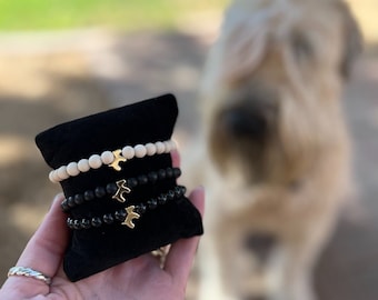 Könnte beinhalten: Drei übereinander gestapelte Armbänder auf einem schwarzen Samtdisplay. Das obere Armband besteht aus hellen Holzperlen, das mittlere Armband aus schwarzen Perlen und das untere Armband aus dunkelgrauen Perlen. Jedes Armband hat einen goldenen Anhänger in Form eines Vogels.