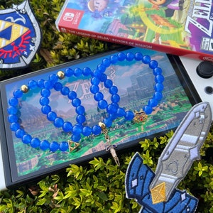 Pode incluir: Um bracelete azul e dourado com tema Zelda com um amuleto da Master Sword. O bracelete está em cima de um console Nintendo Switch com o jogo "The Legend of Zelda: Echoes of Wisdom" no fundo.