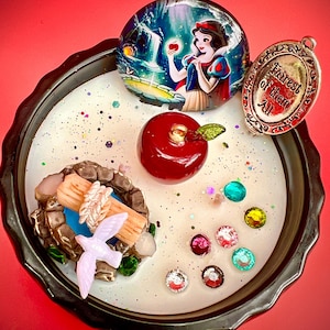 May include: A white candle with a black rim, featuring a miniature scene with a red apple, a bird, a well, and a small glass dome with a picture of Snow White. The candle also includes a silver charm with the words "Fairest of them All".