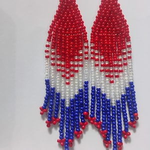 May include: A pair of beaded earrings with a patriotic color scheme. The earrings feature a red, white, and blue pattern, with red beads at the top, white in the middle, and blue at the bottom. The earrings are designed with a fringe style.