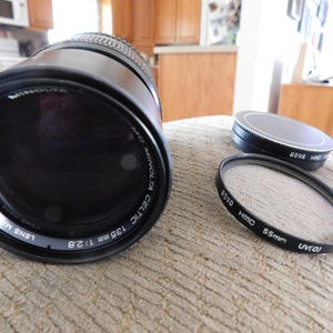 May include: A black Minolta Celtic 135mm 1:2.8 camera lens with a 55mm filter and a lens cap. The lens and filter are black with white lettering. The lens cap is black and silver.