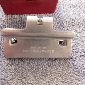 May include: A silver-colored metal tool with the text "PAT PEND BUCK HONEMASTER" is displayed against a textured, light blue background. A red object is partially visible in the background. The tool has a rectangular shape with two holes and a central raised section.