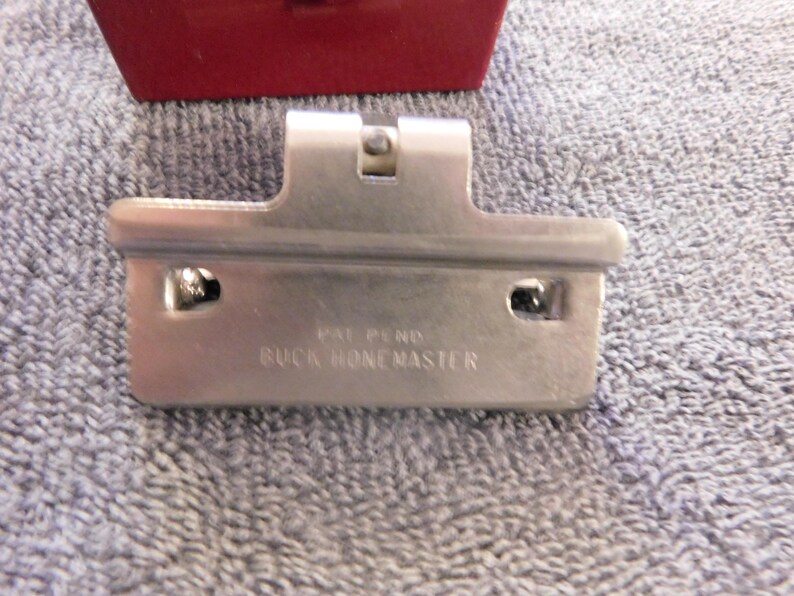 May include: A silver-colored metal tool with the text "PAT PEND BUCK HONEMASTER" is displayed against a textured, light blue background. A red object is partially visible in the background. The tool has a rectangular shape with two holes and a central raised section.