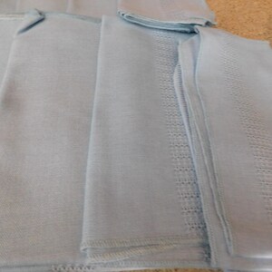 May include: A stack of light blue linen napkins with decorative embroidered borders. The napkins are folded and arranged, showcasing the detailed stitching and soft texture of the fabric. The color is a pale, calming blue.
