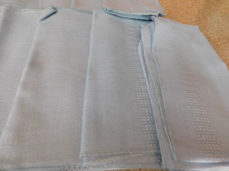 May include: A stack of light blue linen napkins with decorative embroidered borders. The napkins are folded and arranged, showcasing the detailed stitching and soft texture of the fabric. The color is a pale, calming blue.