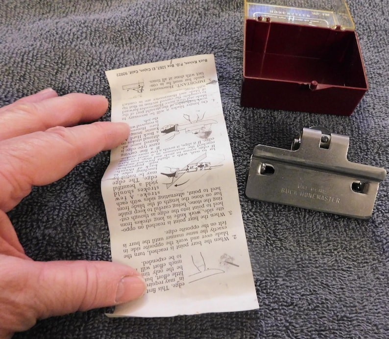May include: A Buck Hone Master knife sharpener with a red and clear plastic case, a metal sharpening guide, and a folded instruction sheet. The instruction sheet has diagrams and text.