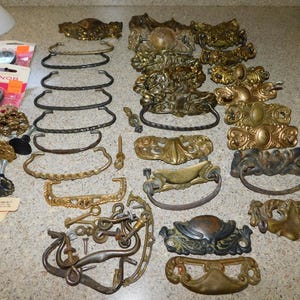 May include: A collection of antique brass and bronze drawer pulls, knobs, and handles. The hardware includes various styles, from ornate floral designs to simple curved handles. Some pieces are still in their original packaging.