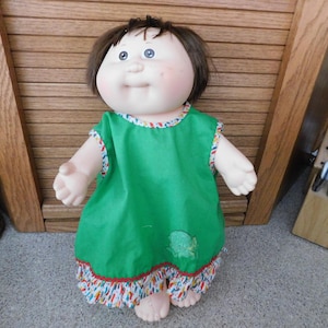 May include: A vintage doll with brown hair and blue eyes, wearing a green dress with colorful trim. The doll has a round face and is standing on a patterned surface.