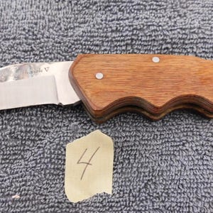 K&ouml;nnte beinhalten: Ein Klappmesser mit einer Klinge aus Edelstahl und einem Holzgriff. Der Griff ist hellbraun und hat ein geschwungenes Design. Das Messer liegt auf einer strukturierten grauen Oberfl&auml;che. Ein kleines St&uuml;ck Papier mit der Zahl 4 liegt neben dem Messer.