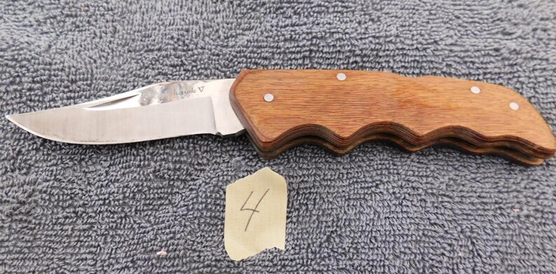 K&ouml;nnte beinhalten: Ein Klappmesser mit einer Klinge aus Edelstahl und einem Holzgriff. Der Griff ist hellbraun und hat ein geschwungenes Design. Das Messer liegt auf einer strukturierten grauen Oberfl&auml;che. Ein kleines St&uuml;ck Papier mit der Zahl 4 liegt neben dem Messer.