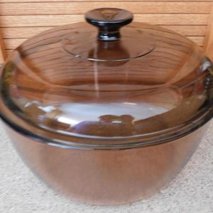 May include: A clear, brown glass casserole dish with a matching lid and a black knob. The dish has two side handles. The casserole dish is sitting on a light-colored surface.