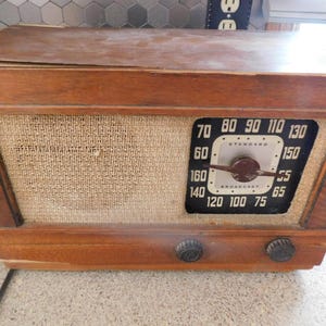 May include: Vintage wooden radio with a woven fabric speaker cover and a black and white dial. The dial displays numbers from 55 to 160, with a central needle. Two dark knobs are located at the bottom.