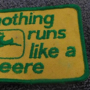 May include: A rectangular yellow fabric patch with a green border and green embroidered text that reads nothing runs like a Deere, featuring a green leaping deer logo, accompanied by a small green and gold diamond-shaped metal pin.