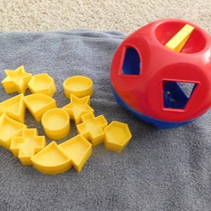 May include: A red and blue plastic shape-sorting toy with a hollow center and a collection of yellow plastic shapes including stars, circles, hexagons, triangles, squares, and crosses that fit into the corresponding openings on the toy's surface.