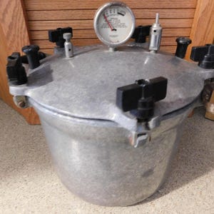 May include: A silver pressure cooker with a gauge and black handles. The cooker has a round body and a lid with multiple knobs and a pressure release valve. The gauge displays pressure readings.