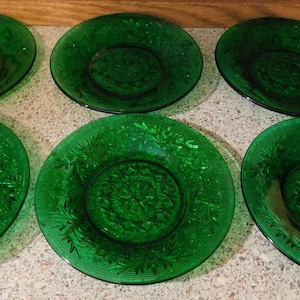 May include: A set of six round, emerald green glass plates arranged in two rows of three. Each plate features a detailed, embossed floral and geometric pattern radiating from the center toward the scalloped edges, creating a textured surface.