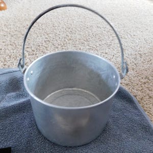 May include: A small, silver metal bucket with a curved handle. The bucket is cylindrical with a slightly flared rim and is sitting on a blue towel. The handle is attached to the bucket with metal hinges.
