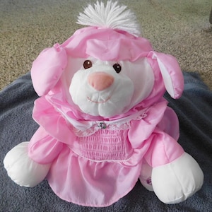 May include: A plush stuffed animal resembling a poodle, featuring white fur on the face and paws, and a pink fabric outfit with a ruffled collar, a smocked bodice, and a white yarn pom-pom on top of its head. The poodle has brown eyes.