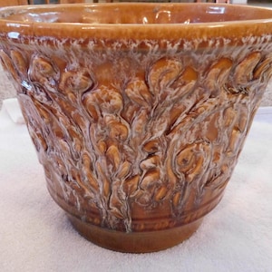 May include: A large, brown ceramic planter with a textured, embossed floral design. The planter has a wide, slightly flared rim and a rounded base. The embossed pattern is accented with white highlights, creating a three-dimensional effect. The planter is suitable for indoor or outdoor use.