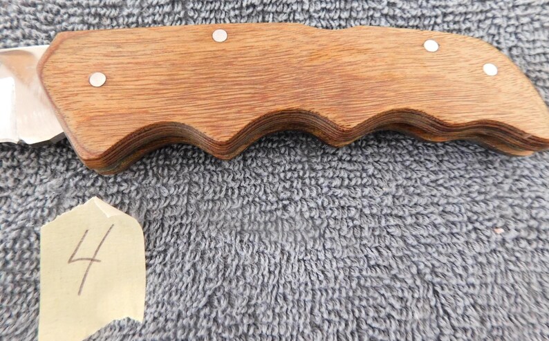 K&ouml;nnte beinhalten: Ein Klappmesser mit einem Holzgriff mit wellenf&ouml;rmigem Design. Der Griff ist mit kleinen silbernen Nieten befestigt. Die Klinge ist teilweise sichtbar, und ein kleines Etikett mit der Nummer "4" befindet sich im Vordergrund.