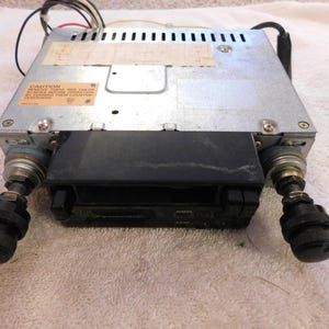 May include: A vintage car stereo with a silver metal casing and a black cassette player. The stereo has two black knobs and a sticker with text. Wires extend from the top.