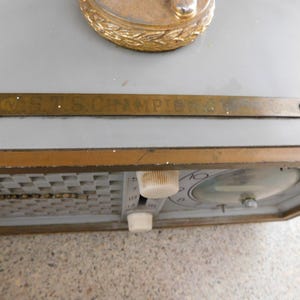 Puede incluir: Un reloj vintage con una esfera gris y un marco dorado. El reloj presenta un patr&oacute;n de cuadros, una esfera redonda y dos perillas blancas. Se ve una placa met&aacute;lica dorada con el texto "C.S.T.S. CHAMPIONS".