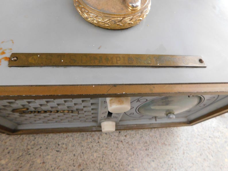 Puede incluir: Un reloj vintage con una esfera gris y un marco dorado. El reloj presenta un patr&oacute;n de cuadros, una esfera redonda y dos perillas blancas. Se ve una placa met&aacute;lica dorada con el texto "C.S.T.S. CHAMPIONS".