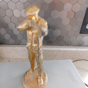 Puede incluir: Estatua dorada de una persona con sombrero y chaqueta, de pie sobre una base redonda. La estatua tiene un aspecto desgastado, con signos visibles de desgaste. El fondo presenta un patr&oacute;n de baldosas hexagonales y un enchufe.