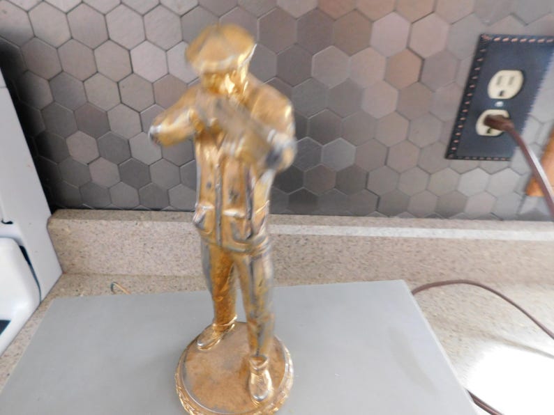 Puede incluir: Estatua dorada de una persona con sombrero y chaqueta, de pie sobre una base redonda. La estatua tiene un aspecto desgastado, con signos visibles de desgaste. El fondo presenta un patr&oacute;n de baldosas hexagonales y un enchufe.