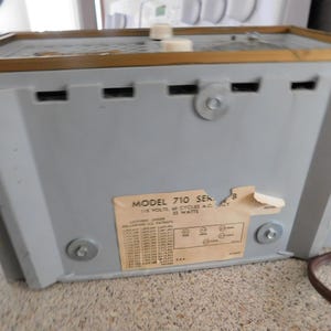Puede incluir: Vista trasera de una radio gris vintage con un borde dorado y un cable de alimentaci&oacute;n marr&oacute;n. La radio tiene una etiqueta con el texto "MODEL 710 SERIES" y especificaciones t&eacute;cnicas. El panel trasero de la radio tiene tornillos y ranuras de ventilaci&oacute;n.