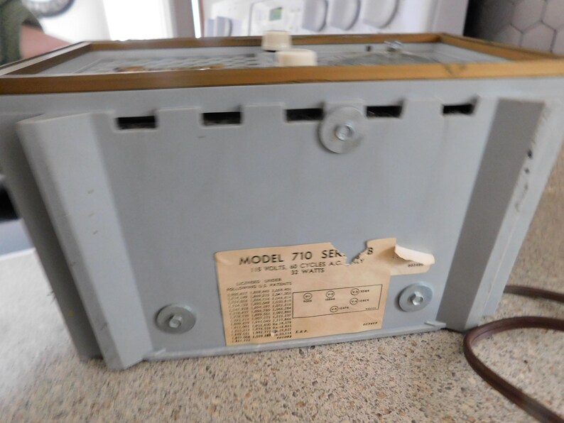Puede incluir: Vista trasera de una radio gris vintage con un borde dorado y un cable de alimentaci&oacute;n marr&oacute;n. La radio tiene una etiqueta con el texto "MODEL 710 SERIES" y especificaciones t&eacute;cnicas. El panel trasero de la radio tiene tornillos y ranuras de ventilaci&oacute;n.