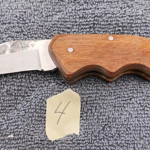 K&ouml;nnte beinhalten: Ein Klappmesser mit polierter Stahlklinge und Holzgriff. Der Griff ist hellbraun und hat ein geschwungenes Design. Die Klinge ist teilweise ge&ouml;ffnet. Ein kleines St&uuml;ck Papier mit der Zahl 4 befindet sich im Bild.
