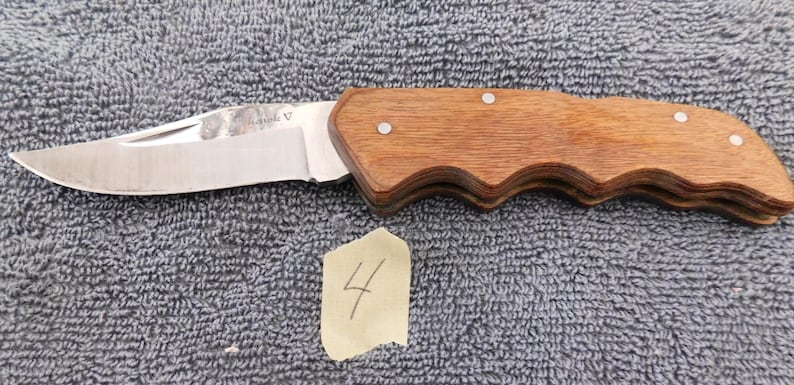 K&ouml;nnte beinhalten: Ein Klappmesser mit polierter Stahlklinge und Holzgriff. Der Griff ist hellbraun und hat ein geschwungenes Design. Die Klinge ist teilweise ge&ouml;ffnet. Ein kleines St&uuml;ck Papier mit der Zahl 4 befindet sich im Bild.