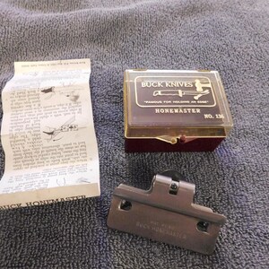 May include: A Buck Knives Honemaster knife sharpener set. The set includes a brown plastic case with the words "BUCK KNIVES" and "HONEMASTER" printed on it, a metal sharpening tool, and a folded instruction sheet.