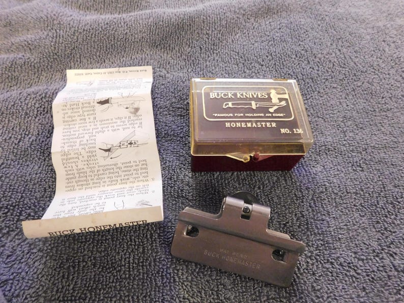 May include: A Buck Knives Honemaster knife sharpener set. The set includes a brown plastic case with the words "BUCK KNIVES" and "HONEMASTER" printed on it, a metal sharpening tool, and a folded instruction sheet.
