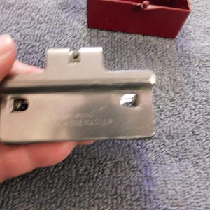 May include: A silver-colored metal tool with the text "ONE MASTER" is held in a hand. The tool has a rectangular shape with a curved edge and two holes. A small red box is visible in the background.