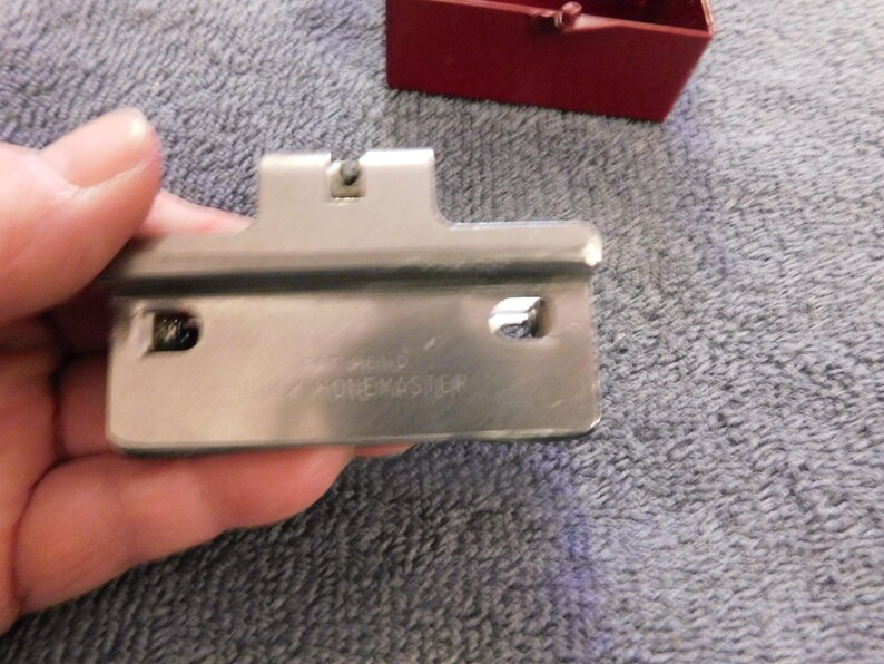 May include: A silver-colored metal tool with the text "ONE MASTER" is held in a hand. The tool has a rectangular shape with a curved edge and two holes. A small red box is visible in the background.